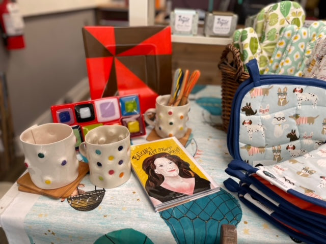 Image is of a display featuring colorful ceramic mugs, a yellow card with an image of Tina Fey, and red and brown fused glass platters. In the background are potholders with dogs printed on them. 