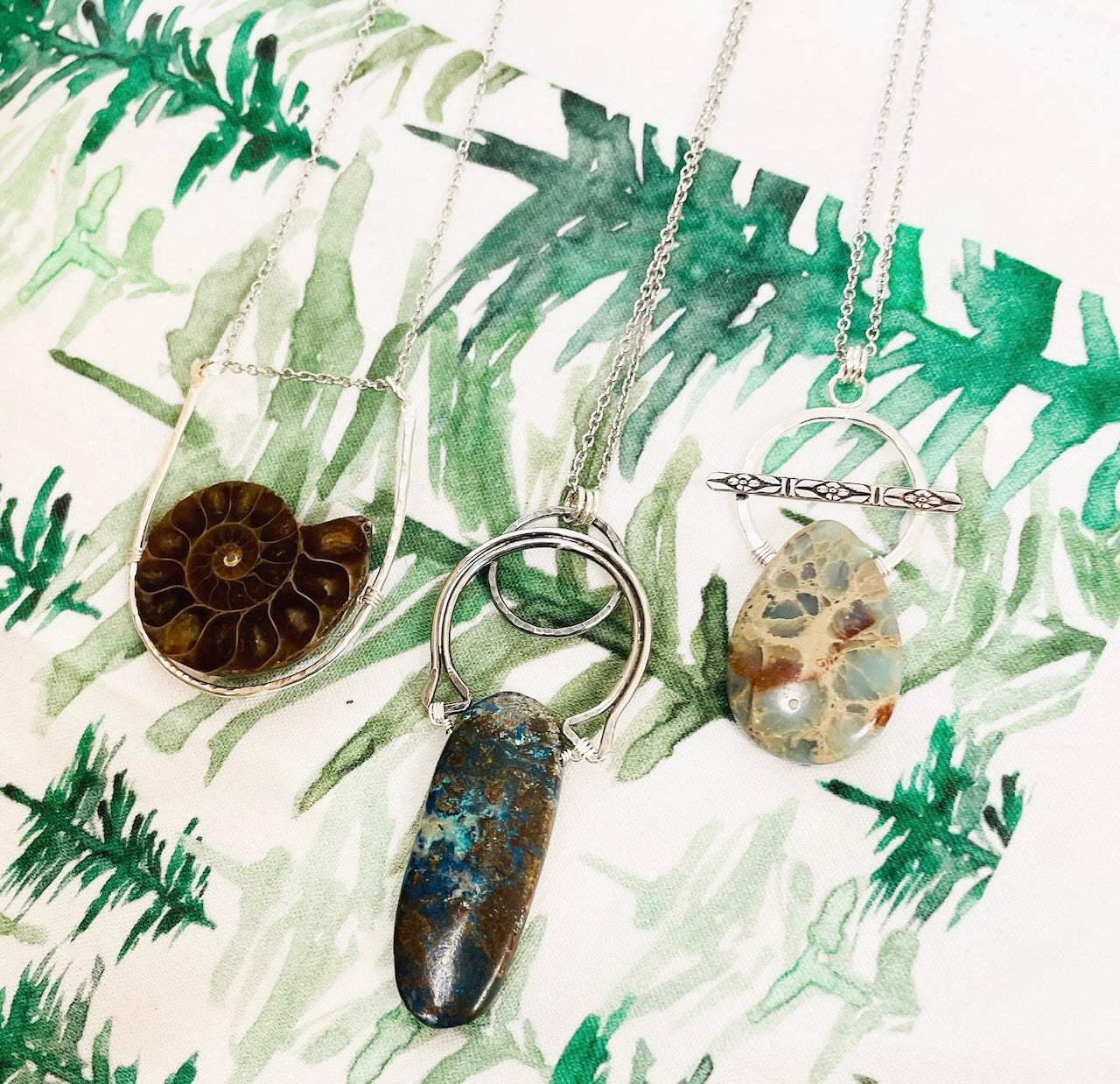 Image shows three silver necklaces, one with a brown ammonite pendant, and two with blue jasper stone pendants. the necklaces lay on a printed cloth background.