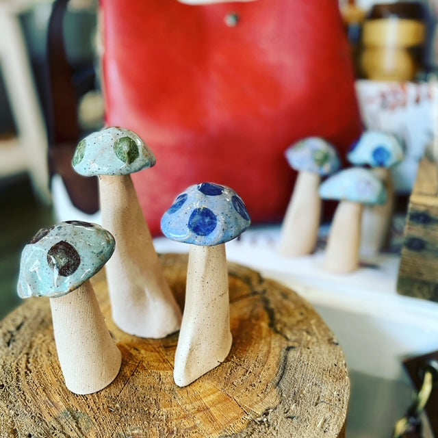 Image shows ceramic mushrooms sitting on top of a wooden stump used as a display. the mushrooms are blue and green. In the background is a red upcycled leather bag. 