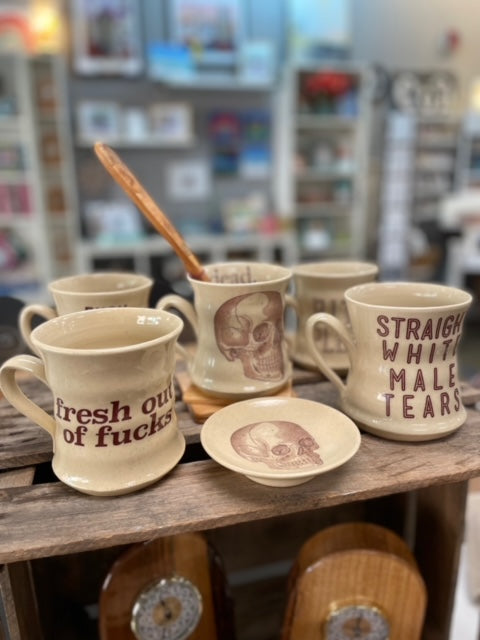 Image shows tan ceramic mugs with phrases such as "fresh out of fucks" and images of skulls printed on them.