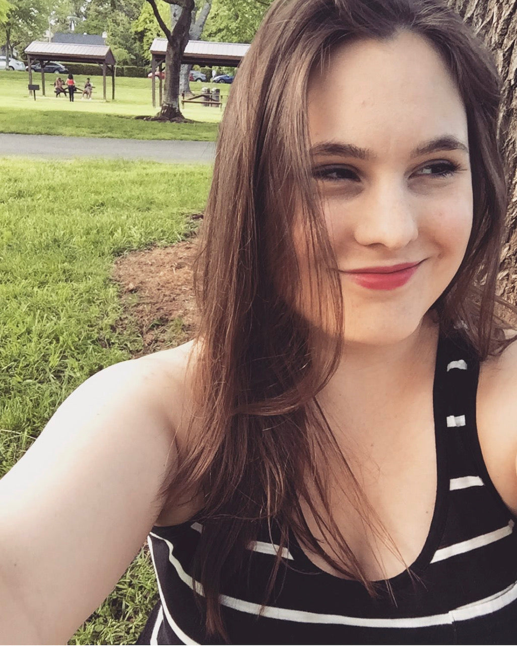 This image shows Muse team member Pam. Pam has long brunette hair and brown eyes. She is sitting in a grassy park and wearing a black and white striped tank top.